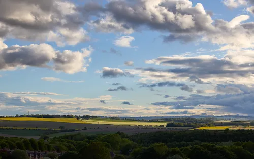 This landscape photograph captures a view across agricultural fields in Derbyshire, England, during a spring evening in the United Kingdom. The main subject of the image is a variety of clouds, ranging from large formations to scattered wisps, set against a clear blue sky above the expansive spring fields. The sunlight bathes the patchwork of fields in warm tones, highlighting the vibrant greenery and the cultivated land typical of agriculture in this region. The photograph effectively showcases the dynamic spring atmosphere in Derbyshire, with the clouds creating a dramatic contrast as they move across the rural landscape.